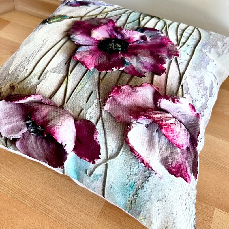 May include: A decorative throw pillow with a white background and a floral design featuring three large pink and purple poppies. The flowers have a textured, painted look.
