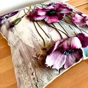 May include: A decorative pillow with a floral design featuring purple poppies on a textured beige background.