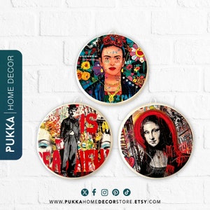 Op de afbeelding: Drie ronde houten wanddecoraties met kleurrijke, abstracte ontwerpen met beroemde kunstenaars zoals Frida Kahlo, Charlie Chaplin en de Mona Lisa. Elk stuk heeft een witte rand en een witte achtergrond met kleurrijke graffiti-achtige ontwerpen.