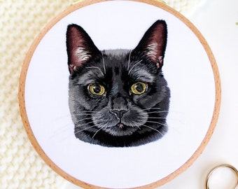 May include: Embroidered black cat portrait in a wooden embroidery hoop. The cat has yellow eyes and is looking directly at the viewer.