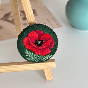 May include: Embroidered round pin featuring a vibrant red poppy flower with a black center and green leaves. The poppy is set against a dark green background with a black border. The pin is displayed on a small wooden easel.