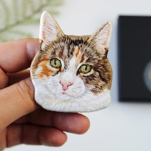 以下が含まれることがあります： 手の中に、茶色、白、オレンジ色の刺繍で猫の顔のパッチを持った手のアップ。猫は緑色の目とピンク色の鼻をしています。
