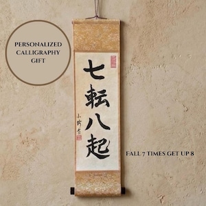 May include: A vertical wall hanging scroll featuring black Japanese calligraphy characters on a cream-colored background. The scroll has gold-toned patterned fabric borders at the top and bottom, with a dark wooden rod at the base and a hanging cord.