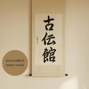 May include: A vertical wall hanging scroll featuring black ink calligraphy characters on a cream-colored paper background. The scroll has dark wooden rods at the top and bottom. A small red seal is visible to the left of the main black text.
