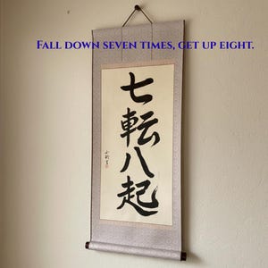 May include: A vertical wall hanging scroll featuring black Japanese calligraphy characters on a cream-colored background. The text reads FALL DOWN SEVEN TIMES, GET UP EIGHT. The scroll has a patterned light gray border and dark wooden end rods.