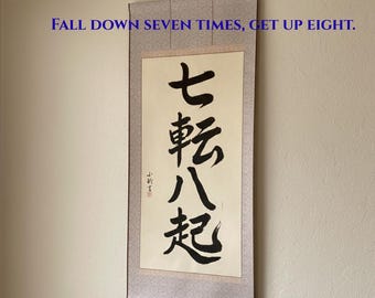 May include: A vertical wall hanging scroll featuring black Japanese calligraphy characters on a cream-colored background. The text reads FALL DOWN SEVEN TIMES, GET UP EIGHT. The scroll has a patterned light gray border and dark wooden end rods.