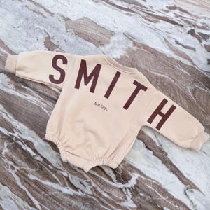May include: A beige baby romper with the name "SMITH" printed in dark brown across the back and sleeves. The word "baby" is printed below the name. The romper is made of a soft material.