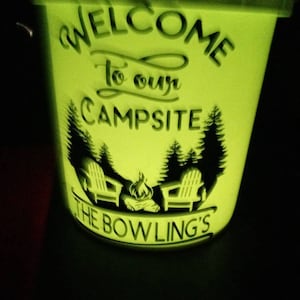 May include: A glowing green bucket with the words "WELCOME To our CAMPSITE" and "THE BOWLING'S" etched into the side. The design includes silhouettes of trees, two chairs, and a campfire.