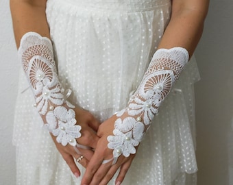 May include: A white lace wedding dress with a pearl necklace and white lace gloves. The dress has a sweetheart neckline and a fitted bodice. The gloves are elbow length and have a delicate floral design.