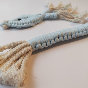 Puede incluir: Un juguete de macramé en forma de pez de color azul claro y crema. El pez está hecho de cuerda de algodón trenzada y tiene una cola larga.