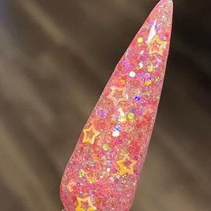 Puede incluir: Una uña rosa con un acabado brillante y detalles en forma de estrella. La uña es de color rosa claro con una capa superior transparente. El brillo es una mezcla de rosa, dorado y colores iridiscentes. Los detalles de estrella son de color dorado claro.