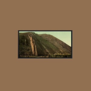 May include: A rectangular framed wall art print featuring a landscape of a steep, grassy hillside with a prominent, light-colored vertical rock formation on the left side, set against a soft, muted sky and a solid brown border background.