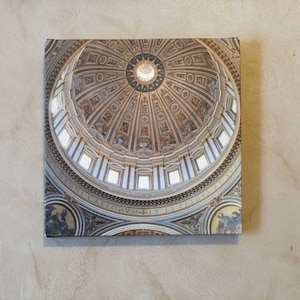 Op de afbeelding: Een close-up van een koepelvormig plafond met ingewikkelde gouden en witte details. Het plafond is versierd met schilderijen van figuren en scènes. De tekst "IN  CALORVM  TV  ES  PETRVS" is rond de rand van de koepel gegraveerd.