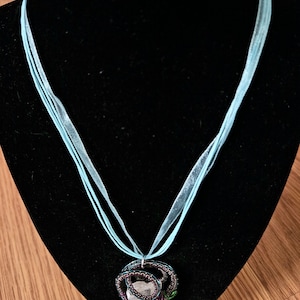 May include: A light blue ribbon necklace with a crystal pendant wrapped in wire. The wire is wrapped in black, purple, and green thread.