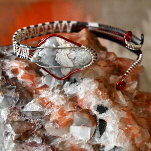Op de afbeelding: Een handgemaakte draadgewikkelde armband met een grijs en wit stenen middelpunt. De armband heeft rode en zilveren accenten, met een zwart-wit geweven patroon op de bovenste band. De sieraden worden getoond op een ruwe, veelkleurige steen.