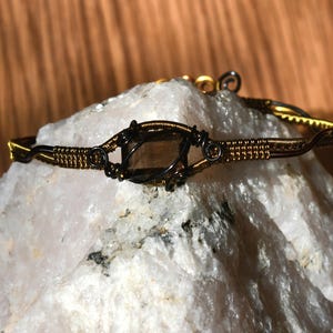 Op de afbeelding: Een met draad omwikkelde armband met een centrale donkere, doorschijnende rookkwarts edelsteen. De band is gemaakt van dunne, gedraaide metaaldraad in een bronskleur, met ingewikkeld metaalwerk en stevige lussen rondom de edelsteen.
