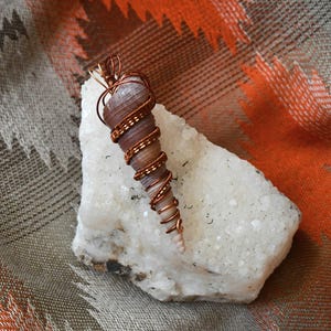 May include: A handcrafted pendant featuring a spiral seashell wrapped in copper wire. The shell is a light brown color, and the wire is a warm copper tone. The pendant rests on a white, textured stone against a patterned fabric background.