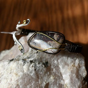 Op de afbeelding: Handgemaakte armband met een grote, patroonsteen in wit, bruin en grijs. De steen is gezet in een zwart en goudkleurig draadwikkel, met zilveren en gouden draaddetails. De armband ligt op een witte rots.