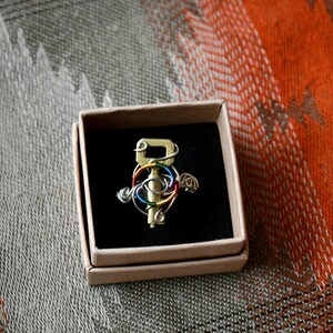 Pode incluir: Um pingente de chave de latão envolto em arame colorido, apresentado em uma pequena caixa de papelão com interior preto. A chave é adornada com arame prateado, vermelho, laranja, amarelo, verde e azul. O fundo apresenta um tecido laranja e cinza padronizado.