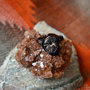 May include: A handcrafted ring with a dark, patterned stone set in a wire wrap of black and copper-colored metal. The ring rests on a rough, textured, brown and white stone, against a blurred orange and gray background.