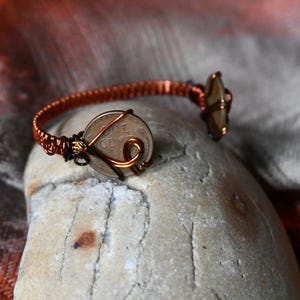Op de afbeelding: Een handgemaakte koperdraadarmband met een Amerikaanse één-centmunt en een decoratieve steen. De armband is gewikkeld in een geweven patroon met een uniek, artistiek ontwerp. De munt is gecentreerd op de armband.