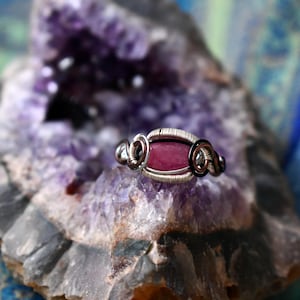 May include: A handcrafted ring with a silver-colored wire design and a rectangular, pink gemstone center. The ring is set against a backdrop of a purple geode, with a blue and purple background. The ring's intricate wirework and the gemstone's color are the focal points.