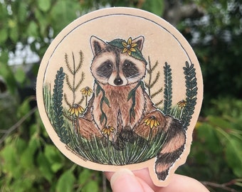 May include: A round sticker featuring a cartoon raccoon wearing a flower crown and sitting in a field of green grass and yellow flowers.