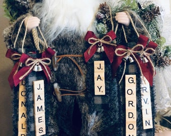 May include: Santa Claus holding four Christmas ornaments made from Scrabble tiles. The ornaments spell out the names "DONALD", "JAMES", "JAY", and "GORDON".