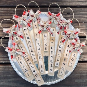 Puede incluir: Adornos navideños hechos a mano. Cada adorno presenta una base de arpillera con letras de Scrabble que deletrean palabras como "Fe", "Amor" y "Alegría". Están adornados con lazos a cuadros rojos y blancos y cuerdas para colgar.