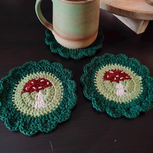 Puede incluir: Tres posavasos de ganchillo con un diseño de hongo. Los posavasos son verdes, verde claro y rojos. Están sobre una superficie de madera oscura.