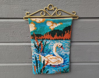 Op de afbeelding: Een witte zwaan geborduurd op een blauw en groen wandkleed met een gouden metalen hanger. Het wandkleed toont een scène van een meer met riet en zonsondergang.