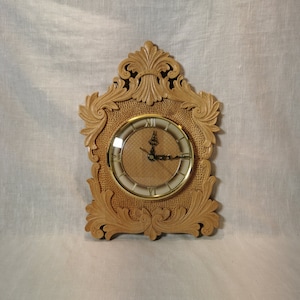 May include: A wooden clock with intricate carvings around the face. The clock face is white with black Roman numerals and gold hands.