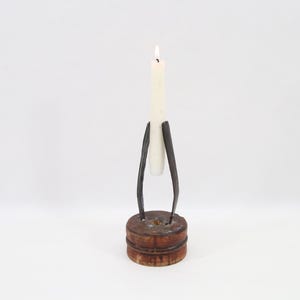May include: A rustic candle holder with a lit, off-white candle. The holder has a dark metal frame that supports the candle, set on a round, wooden base. The design is simple and elegant, suitable for home decor.