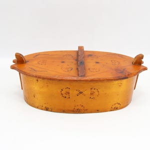 Op de afbeelding: Ovale, antieke houten kist met deksel en handvat. De kist is lichtbruin met donkerbruine accenten en decoratieve patronen. Het deksel heeft een handvat en twee kleine, decoratieve uitsteeksels aan weerszijden.