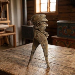 May include: A weathered wooden nutcracker shaped like a person wearing a cap. The figure has a carved face with an open mouth and a long, pointed beard. The wood features a distressed, light brown finish with visible grain and dark markings.