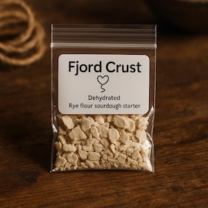 Peut inclure: Un sachet transparent rempli d'un démarreur de levain déshydraté à la farine de seigle. L'étiquette blanche porte l'inscription "Fjord Crust" sur un symbole de cœur, avec "Déshydraté Levain de farine de seigle" en dessous. Le contenu est beige et granuleux.
