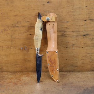 May include: A hunting knife with a fur-covered handle and a leather sheath. The knife has a silver bolster and a dark blade. The sheath is tan with a decorative image and metal rivets.