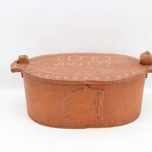 May include: A rustic, oval-shaped wooden box in a warm orange hue. The lid features faded white lettering and is adorned with small, decorative rivets. Wooden knobs are on each end. A vintage storage container.