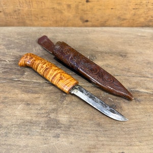 Peut inclure: Un couteau artisanal avec un manche en bois clair et une lame argentée, accompagné d'un étui en cuir marron. L'étui présente des motifs sculptés complexes. Le couteau et l'étui sont posés sur une surface en bois.
