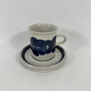 May include: A white ceramic coffee cup and saucer with a blue floral design. The cup has a handle and the saucer has a blue rim.