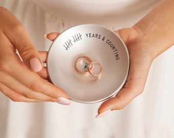 Puede incluir: Un joyero de plata con el texto "Years & Counting" y marcas de hash. Dos anillos de oro descansan en el joyero.
