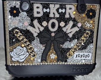 Op de afbeelding: Een zwarte tote bag met de tekst B-K-R M-O-M in strass-letters. De tas is versierd met gouden kettingschakels, een grote zwarte stoffen strik, een witte rozenpatch, zwarte bloemaccenten en een wit rechthoekig label met de tekst XOXOXO.