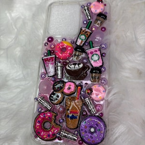May include: A clear phone case with a pink, purple, and white background. The case is decorated with various 3D charms, including donuts, coffee cups, and straws. The charms are in pink, purple, brown, and white.