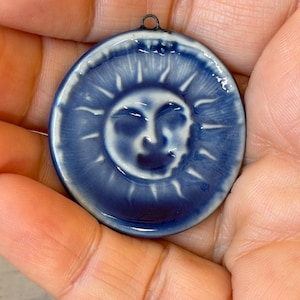 May include: A round, blue and white ceramic pendant featuring a smiling sun face. The pendant has a dark blue border and a small metal loop at the top for hanging. The sun's face is white with blue details.