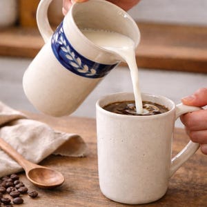 Op de afbeelding: Een crèmekleurige keramische kan met een blauwe bloemenband giet melk in een bijpassende mok met koffie. Een houten lepel, koffiebonen en een linnen servet liggen op een houten oppervlak.
