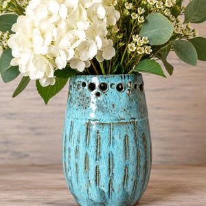 May include: A turquoise ceramic vase with a speckled finish and vertical carved indentations. The top rim features a series of circular cutouts. The vase is filled with a bouquet of white hydrangea flowers and green eucalyptus leaves.