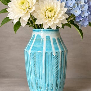 May include: A turquoise ceramic vase with a white drip glaze finish and vertical carved patterns, filled with a bouquet of white dahlias, blue hydrangeas, and small white daisies with green leaves.