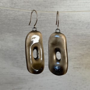 May include: A pair of bronze-colored, oblong earrings with a central oval cutout. The earrings hang from simple, curved ear wires. The metal has a slightly textured surface and a reflective quality, catching the light.
