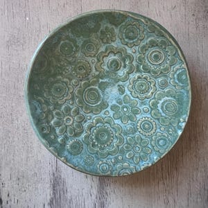 May include: A small, round, green ceramic dish with a floral pattern. The dish has a raised design and a slightly textured surface.