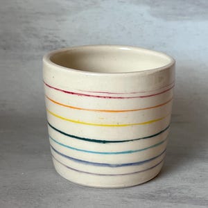 May include: A white ceramic mug with a rainbow stripe design. The stripes are red, orange, yellow, green, blue, and purple.